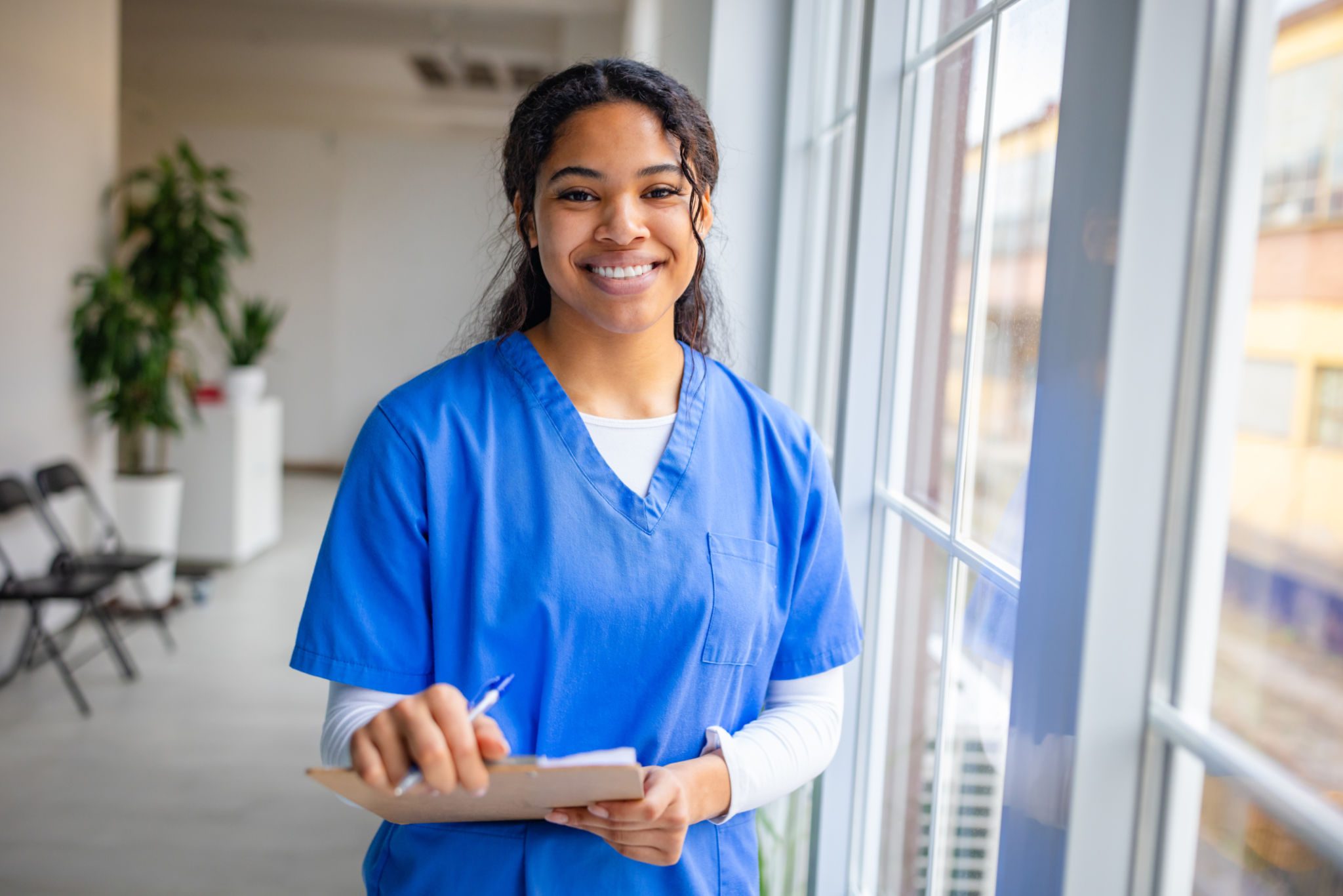 Smiling nurse holding clipboard by window.